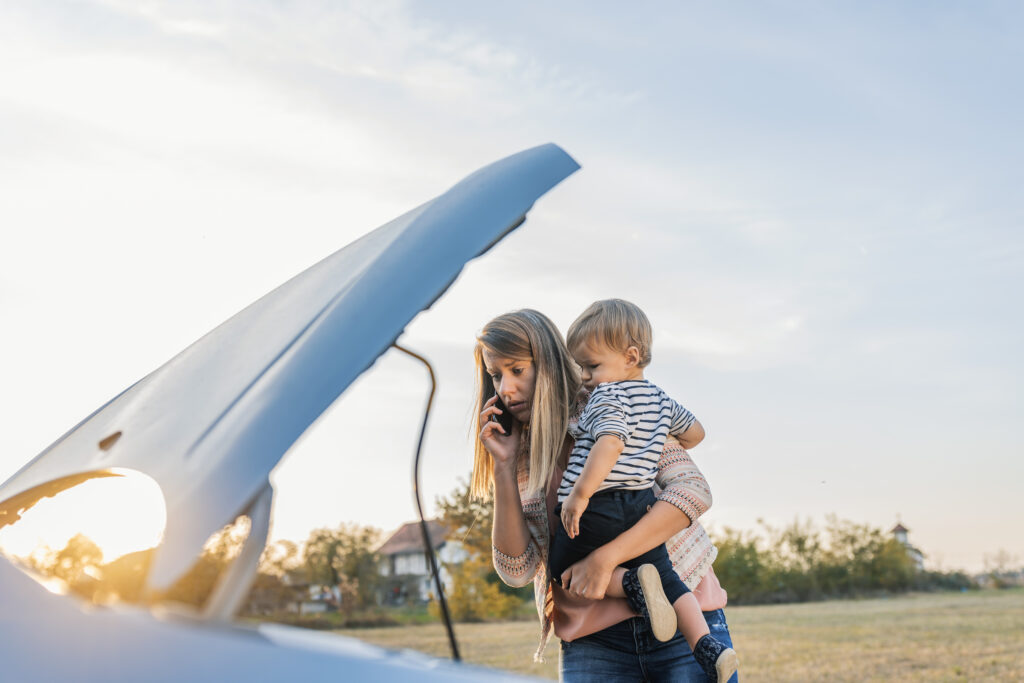 Portrait of young Caucasian woman standing by broken down car and waiting for assistance while holding her baby boy during sunset and talking on cell phone. 